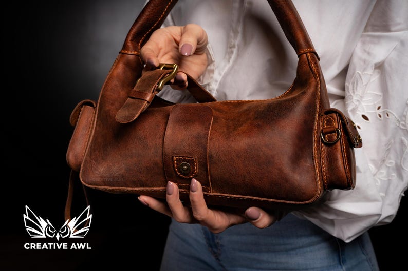 Puede incluir: Un bolso de mano de cuero marr&oacute;n con cierre de solapa y hebilla. El bolso tiene un bolsillo peque&ntilde;o en la parte delantera y un bolsillo m&aacute;s grande en la parte trasera. El bolso est&aacute; sostenido por la mano de una persona.