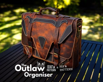 May include: A brown leather briefcase with a black leather flap and a brass buckle. The bag has a handle and a shoulder strap. The text "The Outlaw Organiser" is printed on the front of the bag. The bag is sitting on a wooden table.