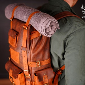 May include: A brown leather backpack with multiple pockets and straps, featuring a rolled-up gray towel secured on top. The backpack has a vintage look with brass buckles and is designed for outdoor adventures.
