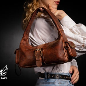 Puede incluir: Un bolso de mano de cuero marr&oacute;n de estilo satchel con cierre de hebilla y un peque&ntilde;o bolsillo en la parte delantera. El bolso tiene un aspecto desgastado y se lleva sobre el hombro.