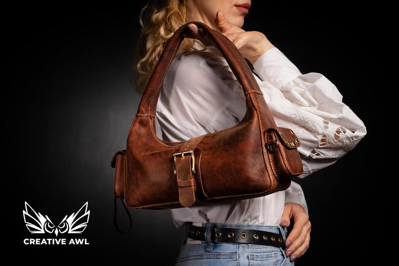 Puede incluir: Un bolso de mano de cuero marr&oacute;n de estilo satchel con cierre de hebilla y un peque&ntilde;o bolsillo en la parte delantera. El bolso tiene un aspecto desgastado y se lleva sobre el hombro.