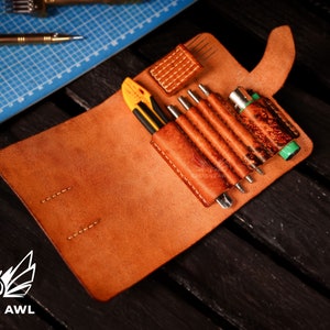 May include: A brown leather tool roll with a flap closure, open to reveal a variety of tools, including a utility knife, awls, and a small screwdriver. The roll is made of distressed leather and has a stitched design. The text "CREATIVE AWl" is visible on the bottom of the image.