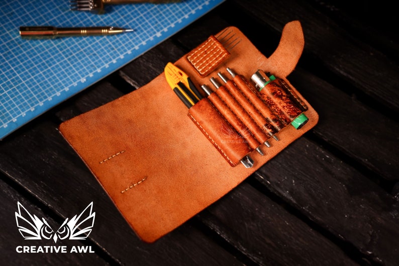 May include: A brown leather tool roll with a flap closure, open to reveal a variety of tools, including a utility knife, awls, and a small screwdriver. The roll is made of distressed leather and has a stitched design. The text "CREATIVE AWl" is visible on the bottom of the image.