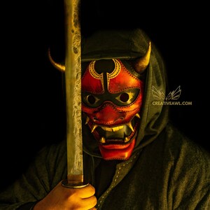 K&ouml;nnte beinhalten: Eine Person tr&auml;gt eine dunkle Kapuzenrobe und eine rote und goldene Oni-Maske mit H&ouml;rnern und h&auml;lt ein Katana-Schwert vor ihrem Gesicht.
