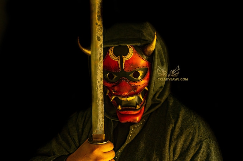 K&ouml;nnte beinhalten: Eine Person tr&auml;gt eine dunkle Kapuzenrobe und eine rote und goldene Oni-Maske mit H&ouml;rnern und h&auml;lt ein Katana-Schwert vor ihrem Gesicht.