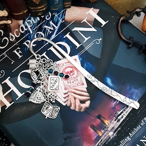 May include: A book titled "Escape from Houdini" by Kerri Maniscalco, a #1 New York Times bestselling author. The book is propped open with a silver bookmark featuring a tarot card design and other charms.