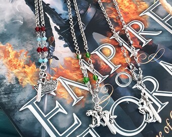 Puede incluir: Cinco cadenas de marcapáginas plateadas con diferentes colgantes. Los colgantes incluyen una corona, un lobo, un grifo, una espada y un dragón. Las cadenas descansan sobre un libro titulado "Empire of Storms, A Throne of Glass Novel" de Sarah J. Maas.