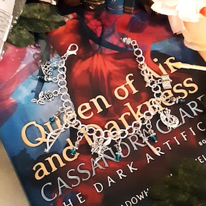 Könnte beinhalten: Ein silbernes Armband mit grünen und silbernen Anhängern, darunter ein Halbmond, eine Fee und ein Hufeisen. Das Armband liegt auf einem Buch mit dem Titel "Queen of Air and Darkness" von Cassandra Clare. Das Buch ist die Fortsetzung des Nr. 1 Bestsellers "The Dark Artifices".