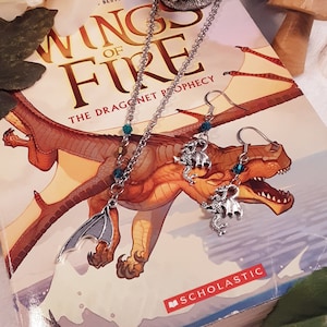 May include: A silver necklace and earrings with dragon charms and teal beads. The jewellery is displayed on a book titled "Wings of Fire: The Dragonet Prophecy" by Tui T. Sutherland.