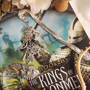 May include: A silver bookmark with a dragon design and green and gold beads hangs from a book titled "The Kings of Clonmel, Book 8" by Flannigan.