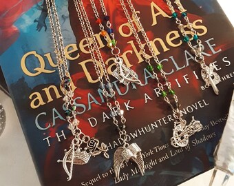 Puede incluir: Un libro titulado "Queen of Air and Darkness" de Cassandra Clare, el tercer libro de la serie Shadowhunters. El libro se muestra con varios collares de plata con dijes inspirados en la serie, incluyendo una corona, alas, un arco y una flecha, y una motocicleta.
