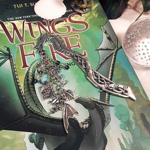 May include: A silver dragon bookmark with intricate details, resting on a book titled "Wings of Fire" by Tui T. Sutherland. The book cover features a green and blue dragon with a black wing.