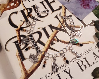 May include: A silver charm bracelet with various charms, including a crown, a castle, a snowflake, and a snake. The bracelet is lying on a book titled "The Cruel Prince" with the text "#1 New York Times Bestselling" and "Holly Black".