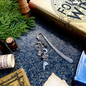 May include: A silver bookmark with a chain and charms, including a dragon, a crescent moon, a castle, and a cloud. The bookmark is lying on a dark surface with a book and a wooden tree ornament.