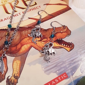 May include: Silver dragon earrings with teal blue crystal accents. The earrings are displayed on a book cover with a dragon illustration and the text "Wings of Fire Dragonet Prophecy Scholastic."