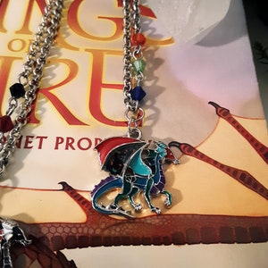 May include: A silver chain necklace with a multi-colored dragon charm. The dragon is made of metal and has red, blue, green, and purple enamel. The necklace is on a book with the words "Wings of Fire" on the cover.