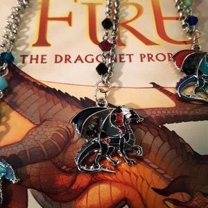 May include: A silver chain necklace with a black, red, and green dragon charm. The dragon has a red heart on its chest and is surrounded by colorful crystals. The necklace is on top of a book titled "Wings of Fire: The Dragonet Prophecy".