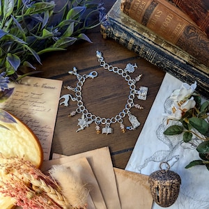 May include: A silver charm bracelet with various charms, including a fairy, a bird, a book, and a camera. The bracelet is displayed on a wooden surface with vintage books, dried flowers, and aged paper. The charms are accented with small, amber-colored beads.