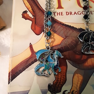 May include: A silver chain necklace with blue and green glass beads and a charm of a blue dragon with wings outstretched. The dragon charm has a detailed design with a light blue body, green wings, and a white belly. The necklace is lying on a book with the title "Wings of Fire: The Dragonet Prophecy" visible.