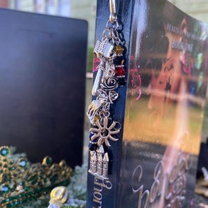 May include: A silver bookmark with charms, including a castle, a sun, a rose, and a sword, hanging from a book titled "A Court of Thorns and Roses" by Sarah J. Maas. The book cover features a red rose and a couple embracing.