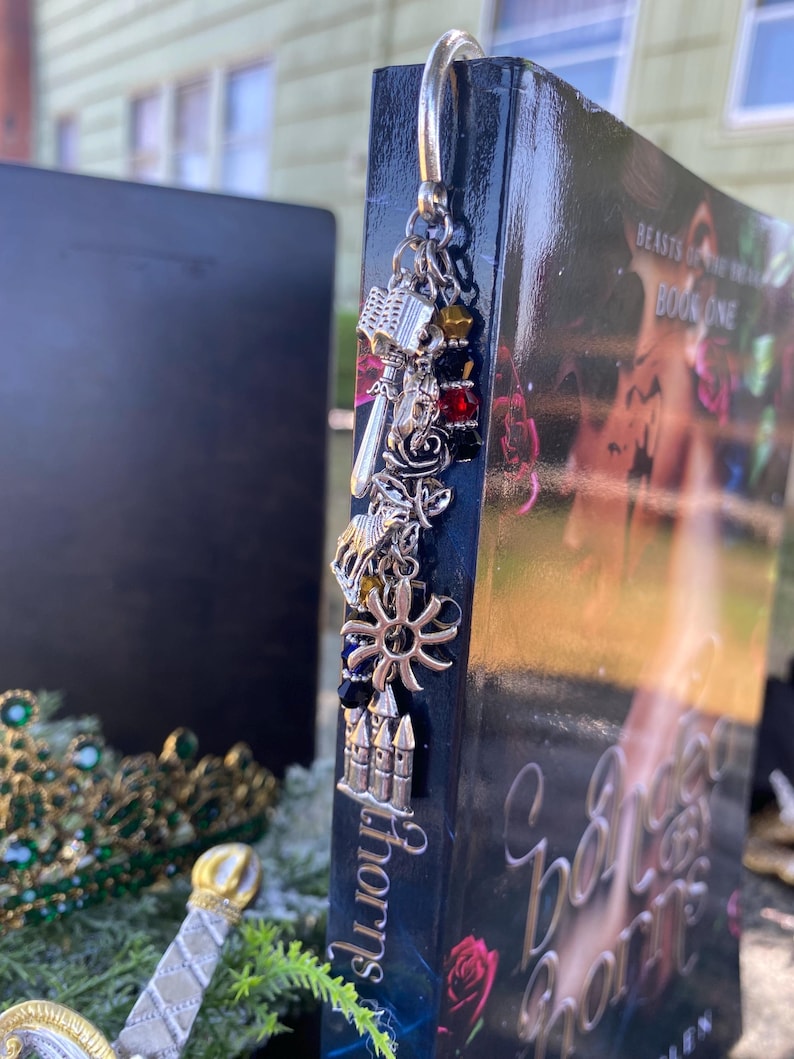 May include: A silver bookmark with charms, including a castle, a sun, a rose, and a sword, hanging from a book titled "A Court of Thorns and Roses" by Sarah J. Maas. The book cover features a red rose and a couple embracing.