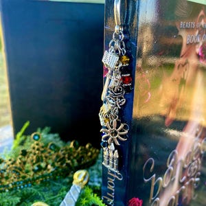 May include: A silver bookmark with charms, including a book, a key, a flower, and a castle, is attached to a book titled "Beast's of the Book One".