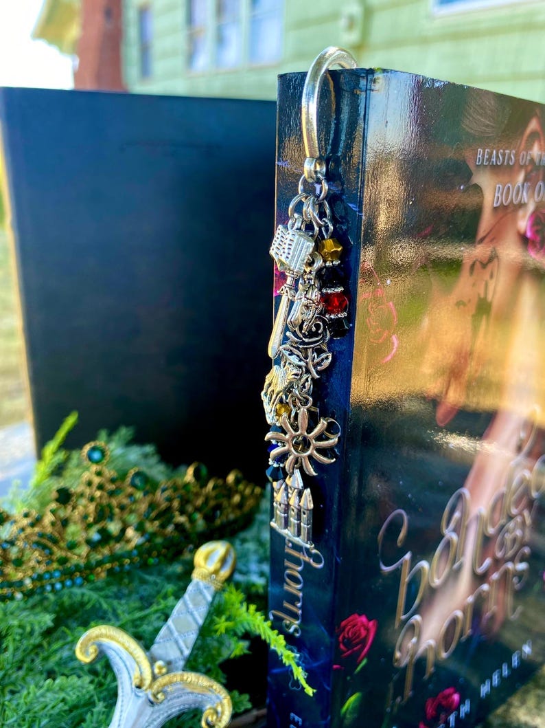 May include: A silver bookmark with charms, including a book, a key, a flower, and a castle, is attached to a book titled "Beast's of the Book One".
