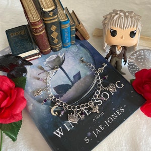 Könnte beinhalten: Ein silbernes Charm-Armband mit violetten Perlen und Charms, darunter ein Halbmond, eine Krone, ein Schlüssel, ein Schloss und ein Vogel. Das Armband liegt auf einem Buch mit dem Titel "Winter Song" von S. Jae-Jones.