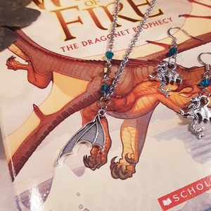 May include: A silver necklace with a dragon wing charm and teal blue beads. The necklace is lying on a book titled "Wings of Fire: The Dragonet Prophecy". Two matching earrings with dragon charms and teal blue beads are also pictured.