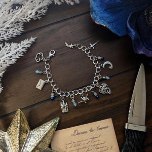 May include: Silver charm bracelet with various pendants, including a cloud, envelope, castle, and crescent moon. The bracelet has blue and clear bead accents. A decorative star and a knife are also visible.