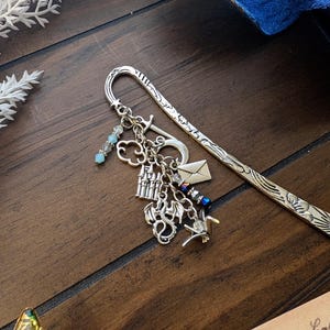 May include: Silver metal bookmark with a decorative etched design and dangling charms. Charms include a cloud, castle, dragon, and envelope. The bookmark has blue and clear crystal beads.