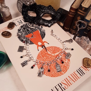 May include: A silver charm bracelet with various charms, including a book, a key, a house, and a crown. The bracelet is lying on a book titled "A Darker Shade of Magic" with a map of London on the cover.