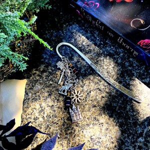 May include: A silver bookmark with a decorative charm featuring a castle, sun, wolf, and other symbols. The bookmark is lying on a textured surface with a book titled "Throned in Thorns" in the background.