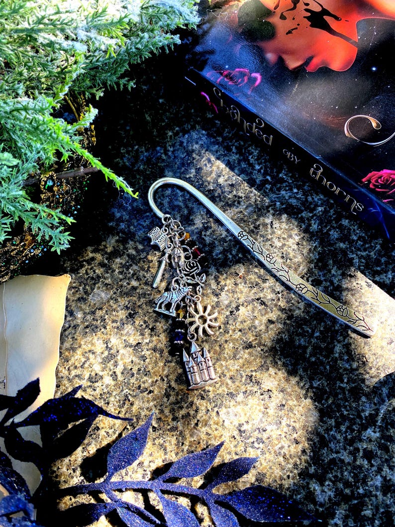 May include: A silver bookmark with a decorative charm featuring a castle, sun, wolf, and other symbols. The bookmark is lying on a textured surface with a book titled "Throned in Thorns" in the background.