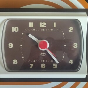 May include: A rectangular brown and white clock with a brown face. The clock face has white numerals and a white and red hand design. The clock is framed in a dark brown border.