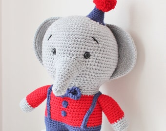 May include: A gray crocheted elephant wearing a red and blue outfit with a red pom-pom hat. The elephant has black eyes and a blue nose.