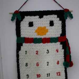 Peut inclure: Un calendrier de l'avent en crochet en forme de pingouin avec un visage de pingouin noir et blanc, une écharpe rouge et verte et les chiffres de 1 à 24.