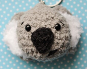 May include: A gray crocheted koala keychain with black eyes and nose, and white fur around the ears. The keychain is attached to a silver ring.