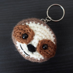 Peut inclure: Un porte-clés en crochet en forme de paresseux avec une fourrure brune et blanche, des yeux noirs et un nez noir. Le porte-clés est attaché à un anneau en métal argenté.
