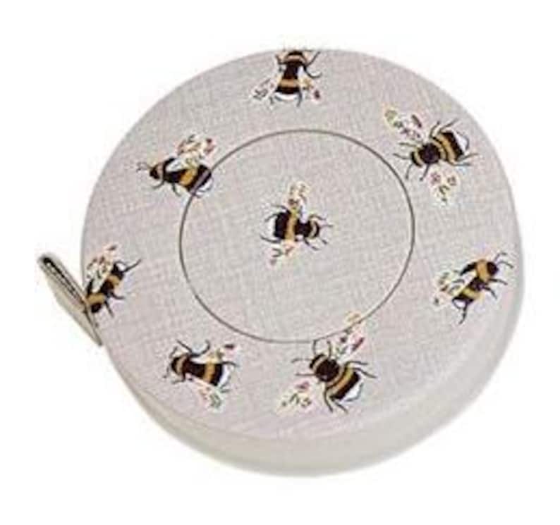 May include: A round, light gray measuring tape with a bee pattern. The tape measure has a white and black bee design with floral accents. The tape measure is in a circular case with a small fabric pull tab.