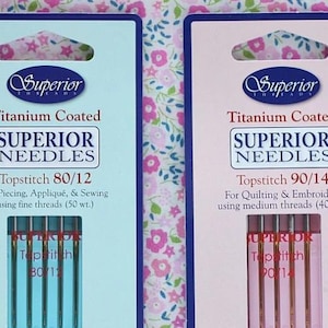 May include: Two packages of Superior Needles are displayed. The packages are labeled "Titanium Coated SUPERIOR NEEDLES." One package is for Topstitch 80/12 needles, and the other is for Topstitch 90/14 needles. The packages are on a floral background.
