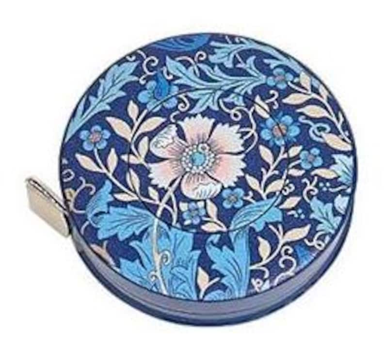 May include: A round, navy blue tape measure with a floral design in shades of blue, pink, and cream. The design features stylized flowers and leaves. A small, silver metal tab extends from the side.