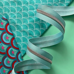 May include: A close-up of fabric and zippers. The fabric has a teal background with a white scale pattern and a red, blue, and teal fan pattern. A teal and white striped zipper with a silver zipper pull is curled in a spiral shape.