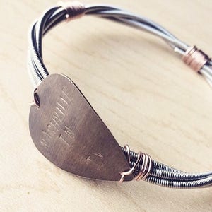 Può includere: Un braccialetto di corde di chitarra argentate con un ciondolo a forma di plettro in rame. Il plettro ha incisa la scritta "Nashville TN".