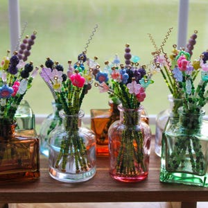 May include: A collection of colorful beaded flower bouquets in various glass vases. The bouquets feature green stems and vibrant flowers in shades of pink, blue, purple, and yellow. The vases are in shades of orange, green, and clear glass.
