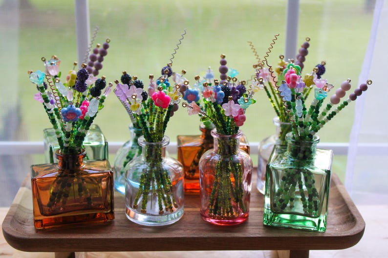 May include: A collection of colorful beaded flower bouquets in various glass vases. The bouquets feature green stems and vibrant flowers in shades of pink, blue, purple, and yellow. The vases are in shades of orange, green, and clear glass.