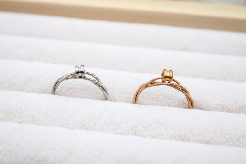 May include: Two guitar string rings, one silver and one gold, displayed in a white velvet ring box.