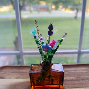 May include: A small, square amber glass vase holding a bouquet of beaded flowers. The flowers are various colors, including blue, pink, and purple, with green beaded stems. The vase sits on a wooden surface near a window.