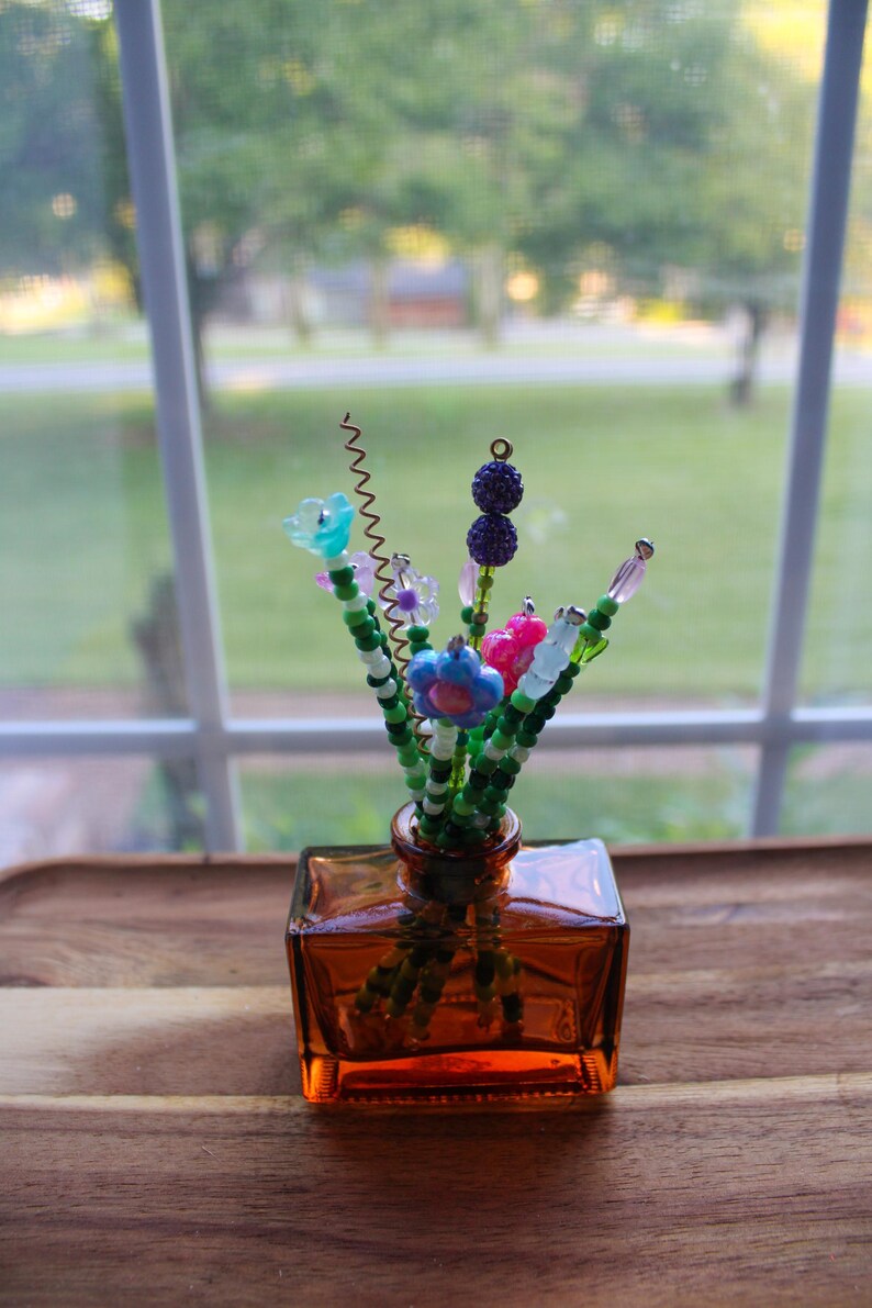 May include: A small, square amber glass vase holding a bouquet of beaded flowers. The flowers are various colors, including blue, pink, and purple, with green beaded stems. The vase sits on a wooden surface near a window.