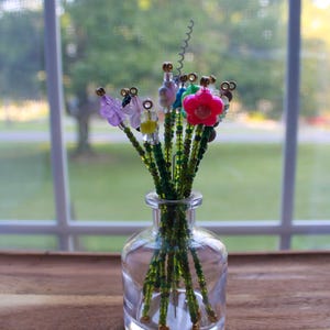 May include: A clear glass vase holding a bouquet of beaded flowers. The flowers have green stems and colorful beaded petals in pink, purple, blue, and yellow. The vase sits on a wooden surface.